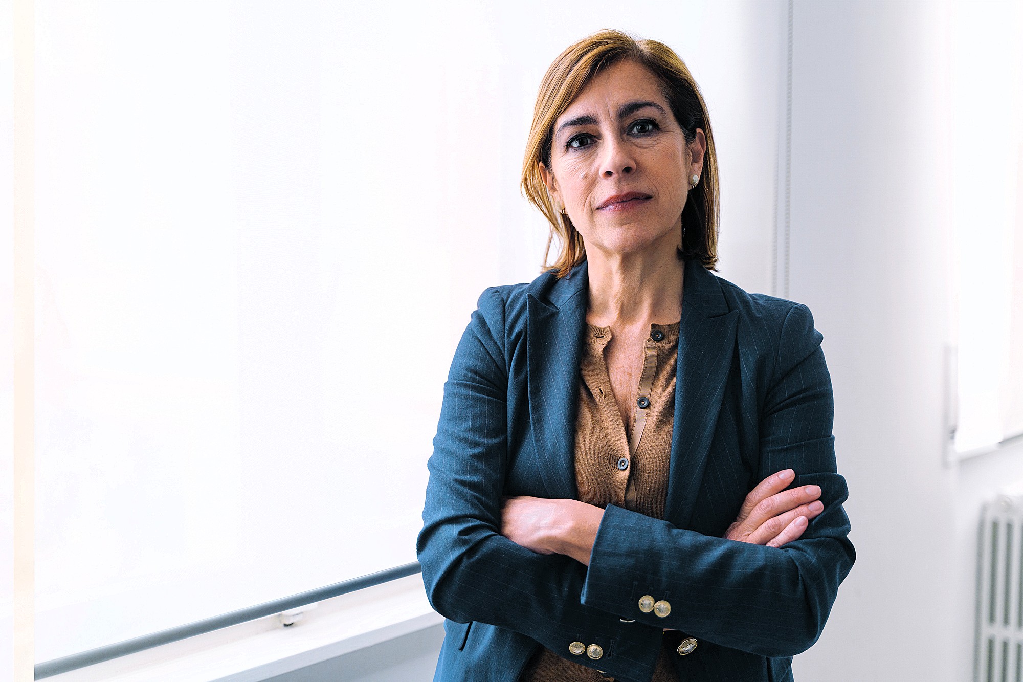 A woman with shoulder-length hair stands confidently in a well-lit room, wearing a dark blazer over a brown top. Her arms are crossed, and she is looking directly at the camera. A large window is visible in the background.