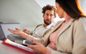 A man and a woman sit on a couch, engaged in a conversation. The man gestures with his hand while looking at the woman, who holds a clipboard and smartphone. They appear to be discussing something seriously.