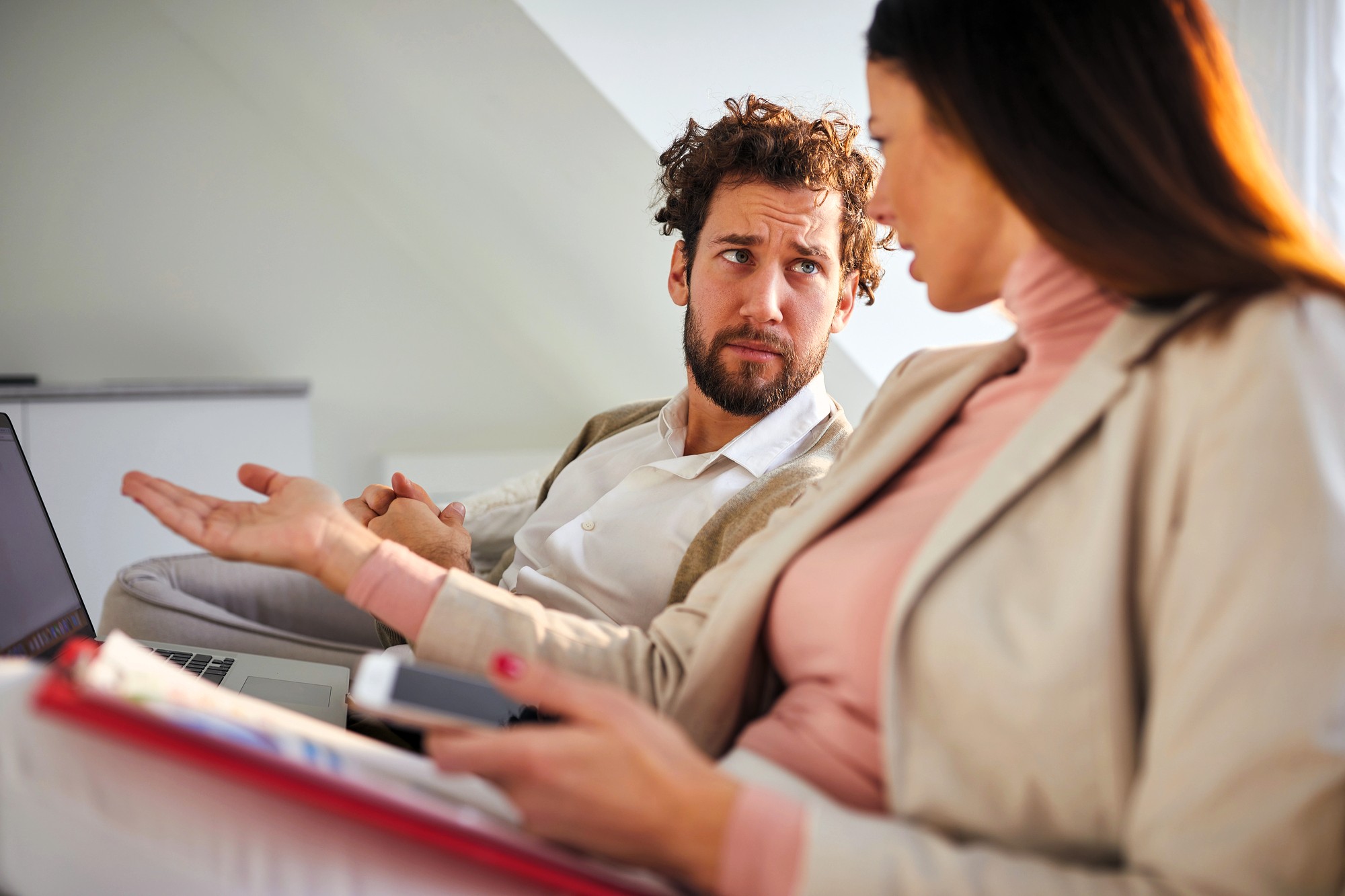 A man and a woman sit on a couch, engaged in a conversation. The man gestures with his hand while looking at the woman, who holds a clipboard and smartphone. They appear to be discussing something seriously.