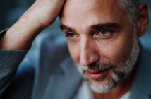Man with short gray hair and beard, wearing a gray suit jacket, looking contemplative. He rests his hand on his head, gazing slightly downwards with a subtle smile. The background is softly blurred.