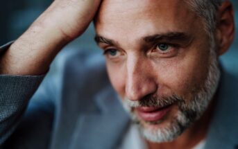 Man with short gray hair and beard, wearing a gray suit jacket, looking contemplative. He rests his hand on his head, gazing slightly downwards with a subtle smile. The background is softly blurred.