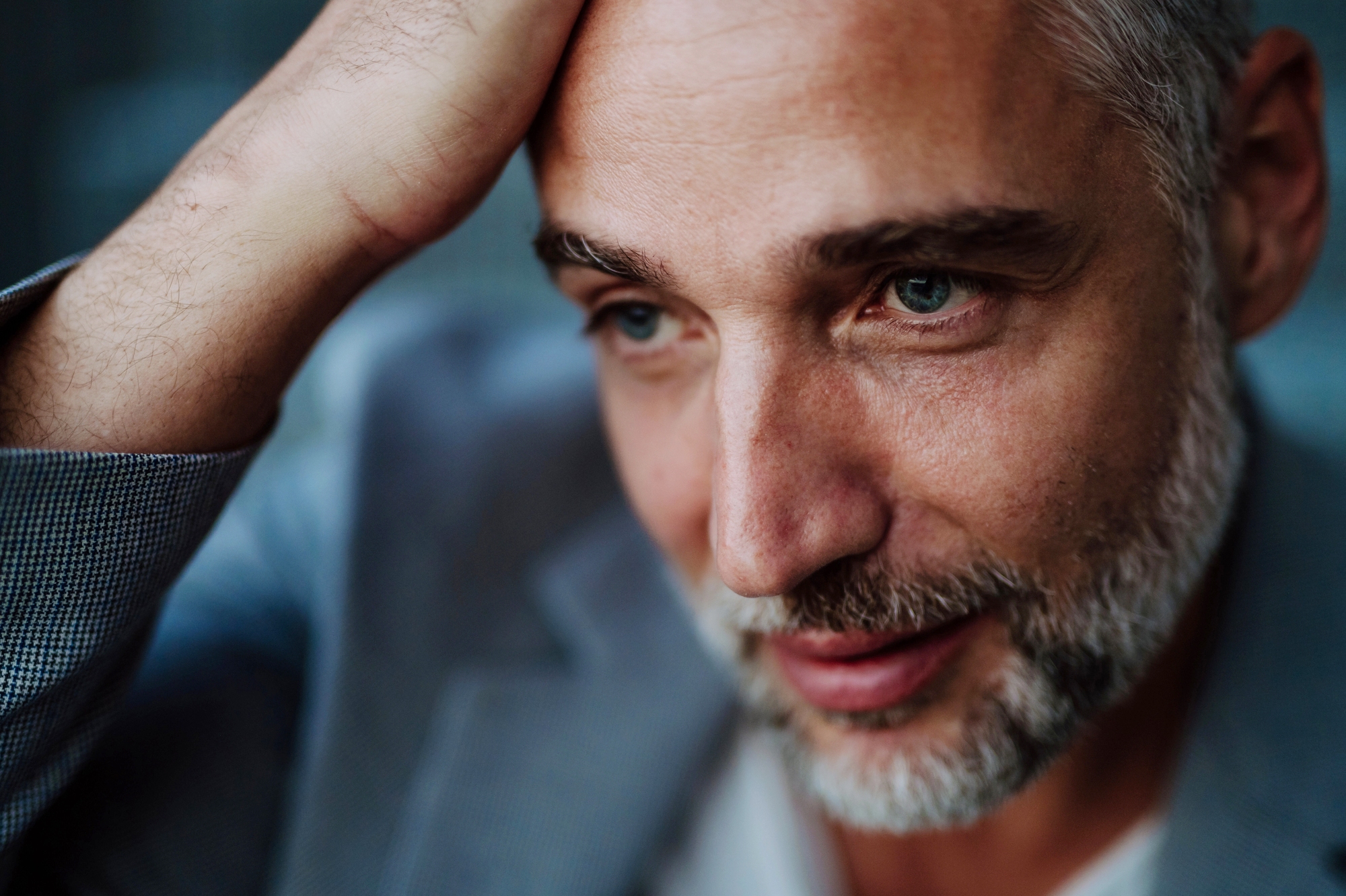 Man with short gray hair and beard, wearing a gray suit jacket, looking contemplative. He rests his hand on his head, gazing slightly downwards with a subtle smile. The background is softly blurred.