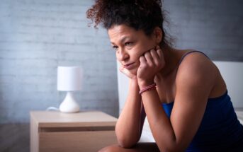 A person with curly hair sits on a bed wearing a blue tank top, resting their chin on their hands and looking thoughtful. A wooden nightstand with a modern white lamp is in the background against a pale brick wall.