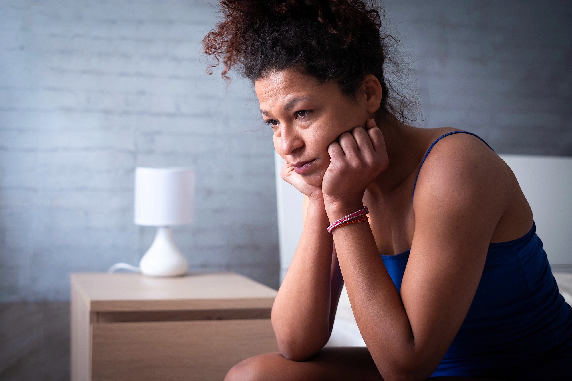 A person with curly hair sits on a bed wearing a blue tank top, resting their chin on their hands and looking thoughtful. A wooden nightstand with a modern white lamp is in the background against a pale brick wall.
