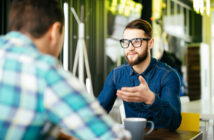 Two people sitting at a table in a modern cafe. One person, with glasses and a beard, gestures while talking. A cup is in front of them. The other person is seen from behind, wearing a checkered shirt. Bright, warm lighting fills the room.