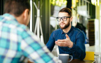 Two people sitting at a table in a modern cafe. One person, with glasses and a beard, gestures while talking. A cup is in front of them. The other person is seen from behind, wearing a checkered shirt. Bright, warm lighting fills the room.