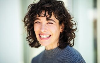 Smiling person with curly dark hair, wearing a gray sweater, standing against a blurred light background.