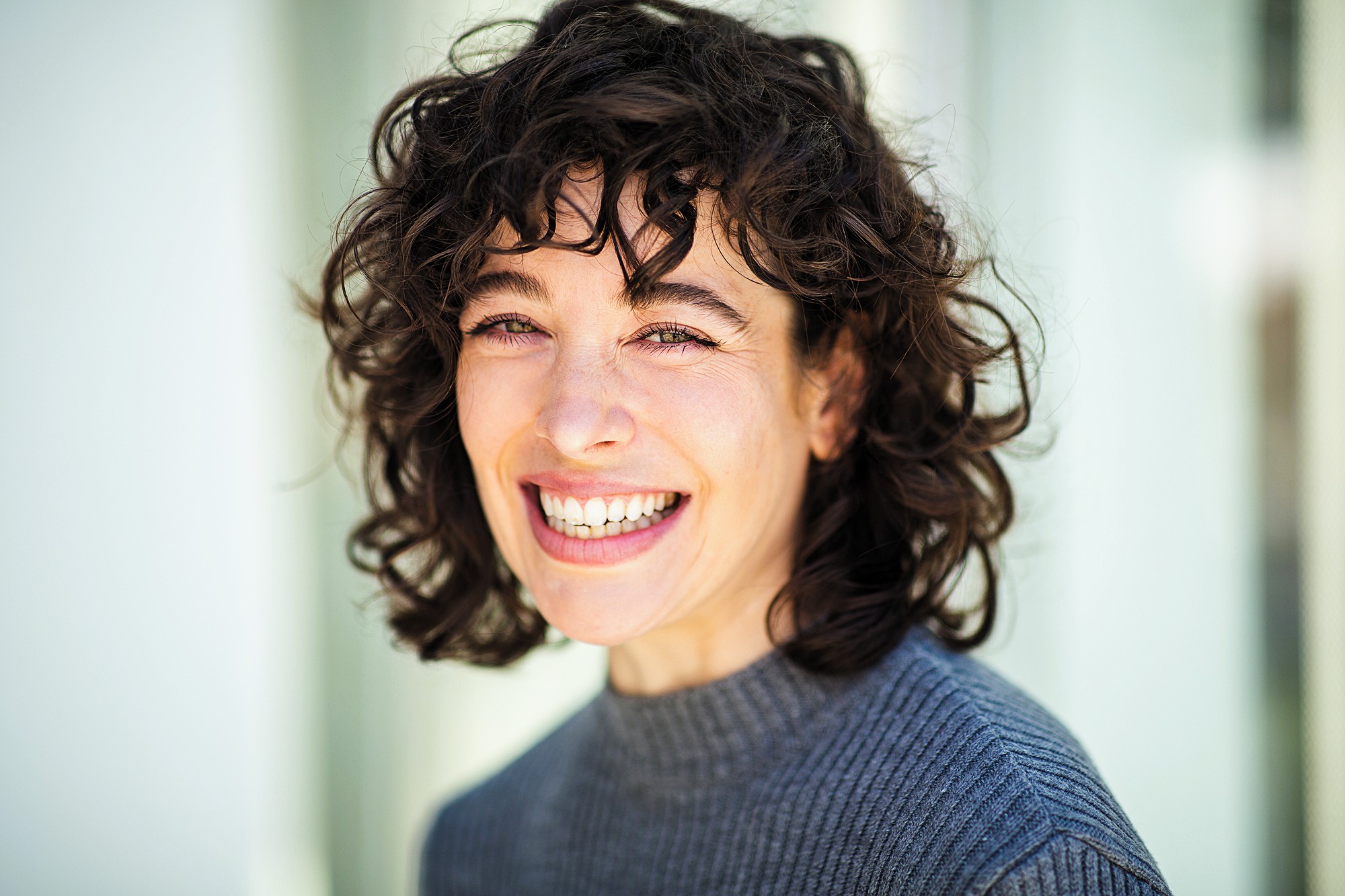 Smiling person with curly dark hair, wearing a gray sweater, standing against a blurred light background.
