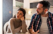 A woman with brown curly hair wearing a beige turtleneck sits at a table, resting her head on her hand, looking away. A man with dark hair and a plaid shirt sits beside her, gesturing as he speaks. They are in a kitchen.