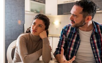 A woman with brown curly hair wearing a beige turtleneck sits at a table, resting her head on her hand, looking away. A man with dark hair and a plaid shirt sits beside her, gesturing as he speaks. They are in a kitchen.