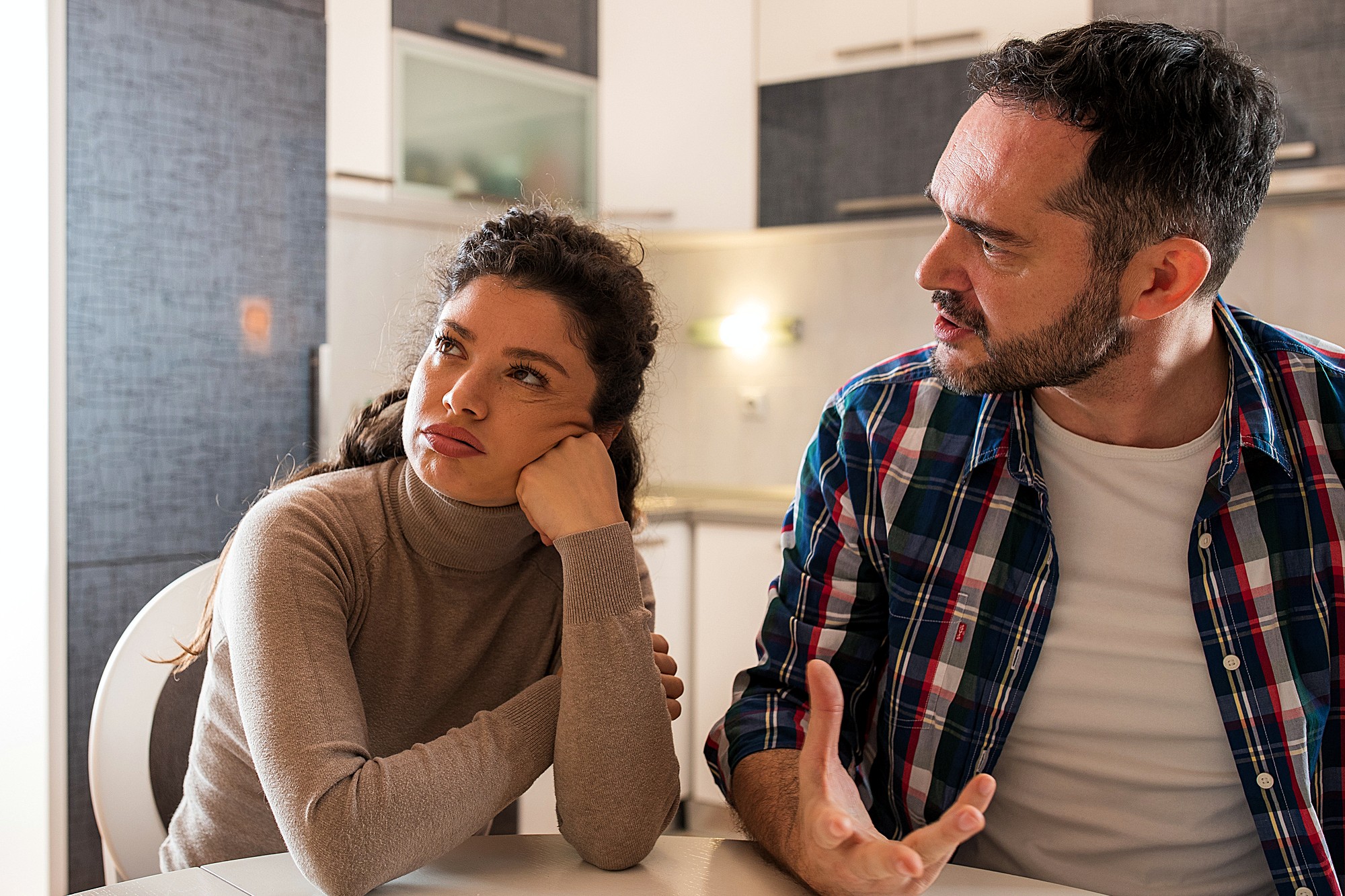 A woman with brown curly hair wearing a beige turtleneck sits at a table, resting her head on her hand, looking away. A man with dark hair and a plaid shirt sits beside her, gesturing as he speaks. They are in a kitchen.