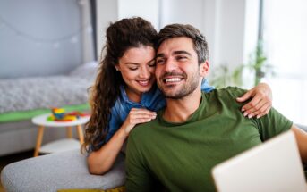A smiling couple sits on a couch in a cozy living room. The woman has her arms around the man, who is holding a tablet. A bed and a small table with toys are visible in the background, surrounded by soft natural light.