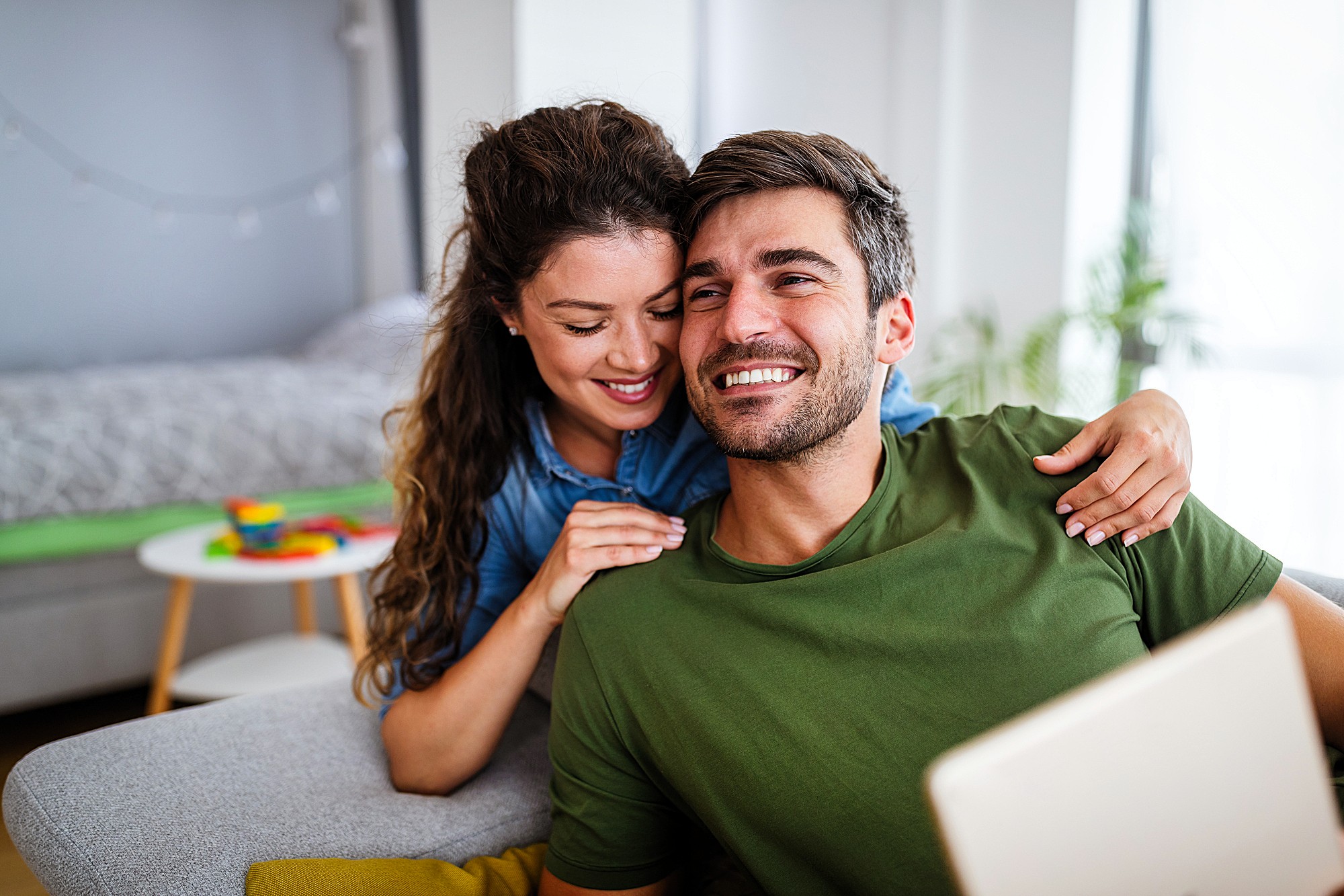 A smiling couple sits on a couch in a cozy living room. The woman has her arms around the man, who is holding a tablet. A bed and a small table with toys are visible in the background, surrounded by soft natural light.