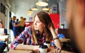 A woman in a plaid shirt sits at a table in a cafe, looking out a window with a thoughtful expression. She holds a cup of coffee. In the blurry background, people are engaged in conversation and activity.