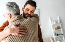 A young man with a beard gently embraces an older man with gray hair, wearing a brown sweater. They appear to be in a living room with white walls and a shelf in the background. The scene conveys a sense of warmth and support.