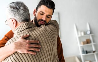 A young man with a beard gently embraces an older man with gray hair, wearing a brown sweater. They appear to be in a living room with white walls and a shelf in the background. The scene conveys a sense of warmth and support.
