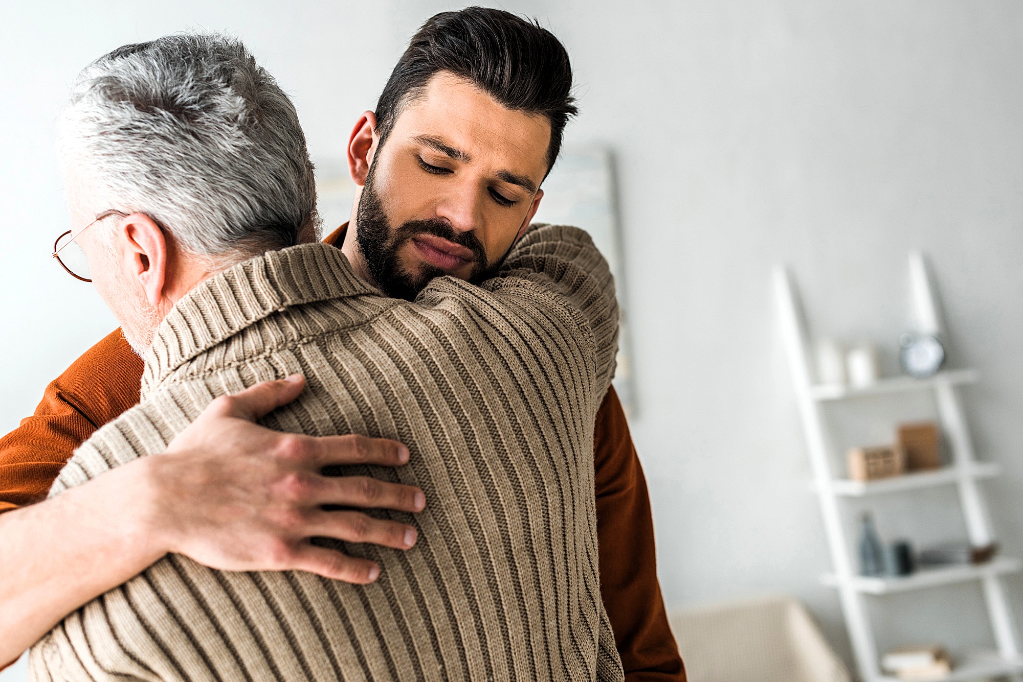 A young man with a beard gently embraces an older man with gray hair, wearing a brown sweater. They appear to be in a living room with white walls and a shelf in the background. The scene conveys a sense of warmth and support.