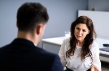 A woman with long dark hair sits at a table, facing a man. She appears to be speaking or listening intently, with a serious or concerned expression. The setting is an office with a blurred background and a minimalist design.