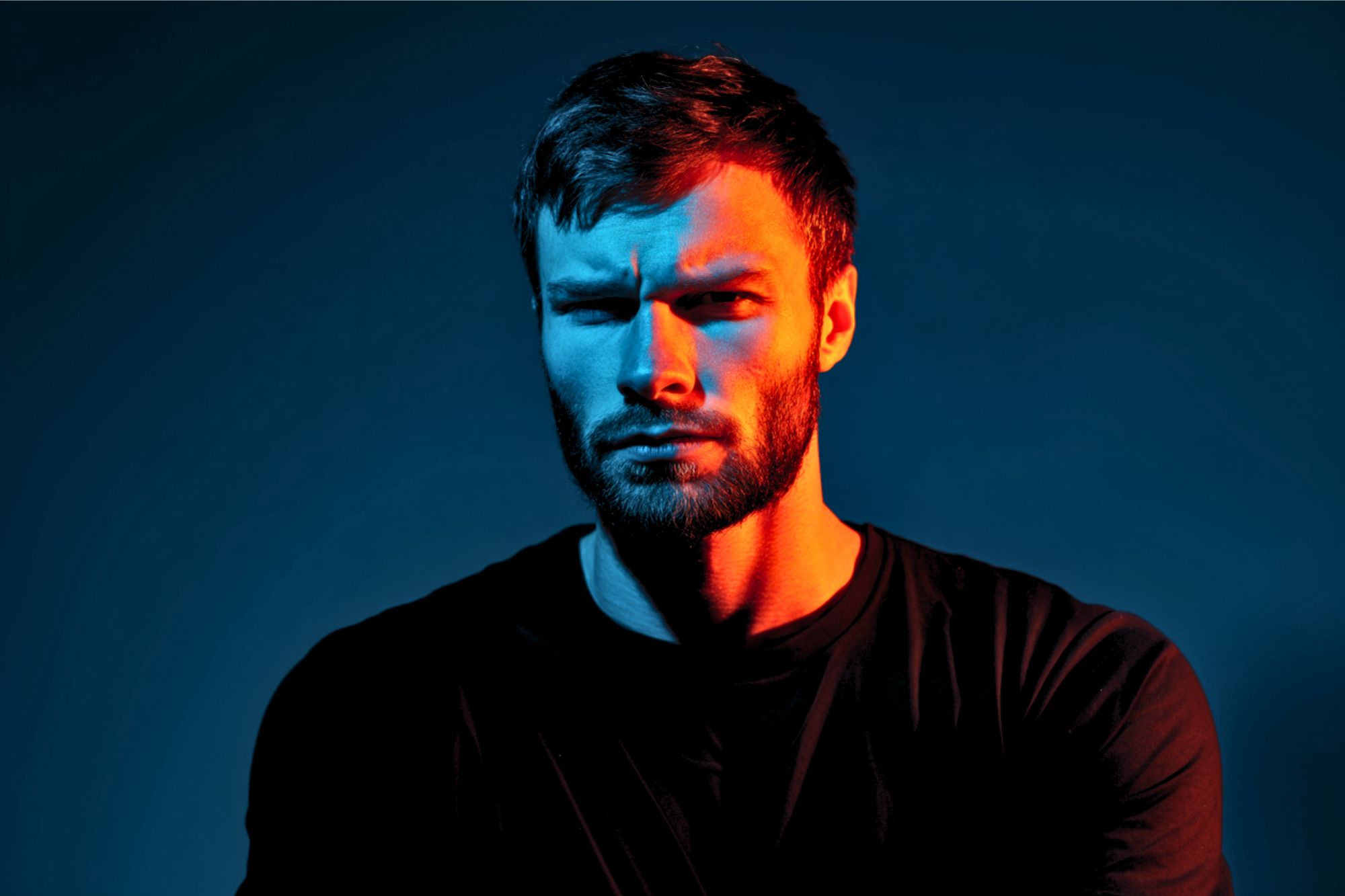 A man with a beard and mustache looks intensely at the camera, lit dramatically with blue and orange lighting on his face, wearing a black shirt against a dark background.