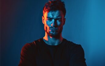 A man with a beard and short hair stands with arms crossed, looking serious. He is lit dramatically with blue and red lighting against a dark background.