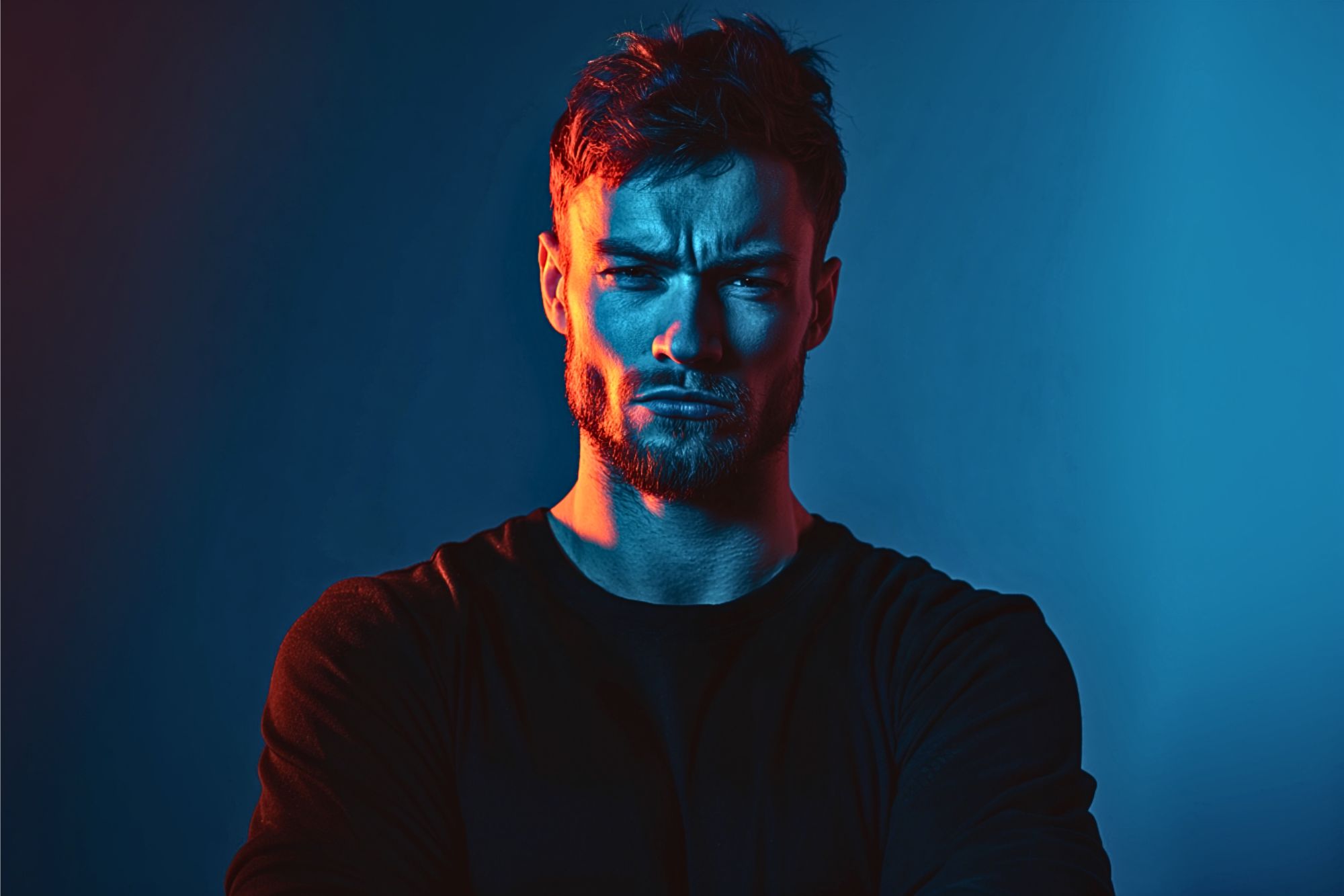 A man with a beard and short hair stands with arms crossed, looking serious. He is lit dramatically with blue and red lighting against a dark background.