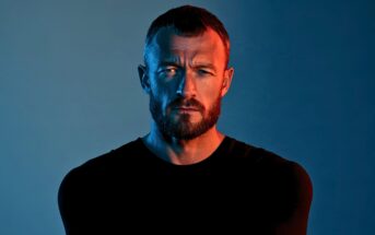 A man with a beard and short hair stares seriously at the camera under dramatic blue and orange lighting, wearing a plain black shirt against a gradient background.