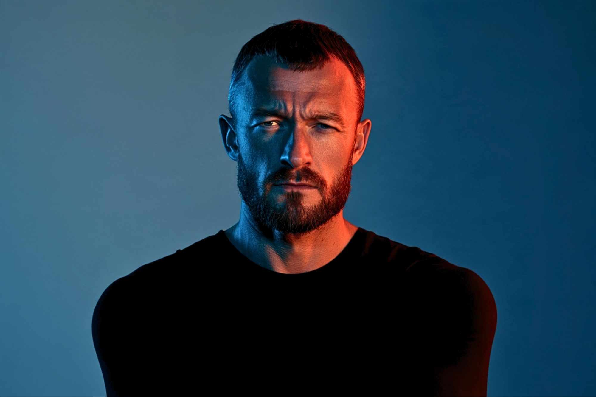A man with a beard and short hair stares seriously at the camera under dramatic blue and orange lighting, wearing a plain black shirt against a gradient background.