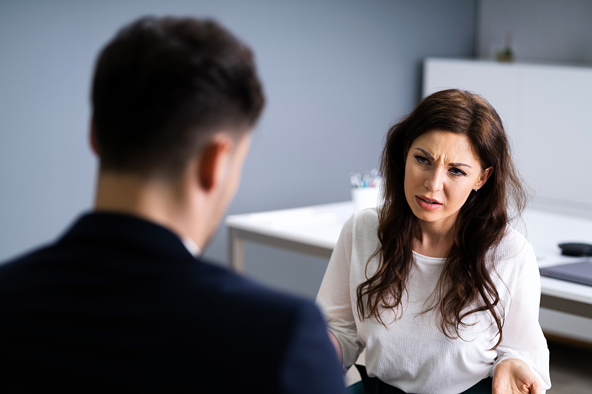 A woman with long dark hair sits at a table, facing a man. She appears to be speaking or listening intently, with a serious or concerned expression. The setting is an office with a blurred background and a minimalist design.