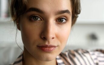 A young person with light brown hair and hazel eyes looking at the camera. They are wearing a striped shirt, and the background is blurred.