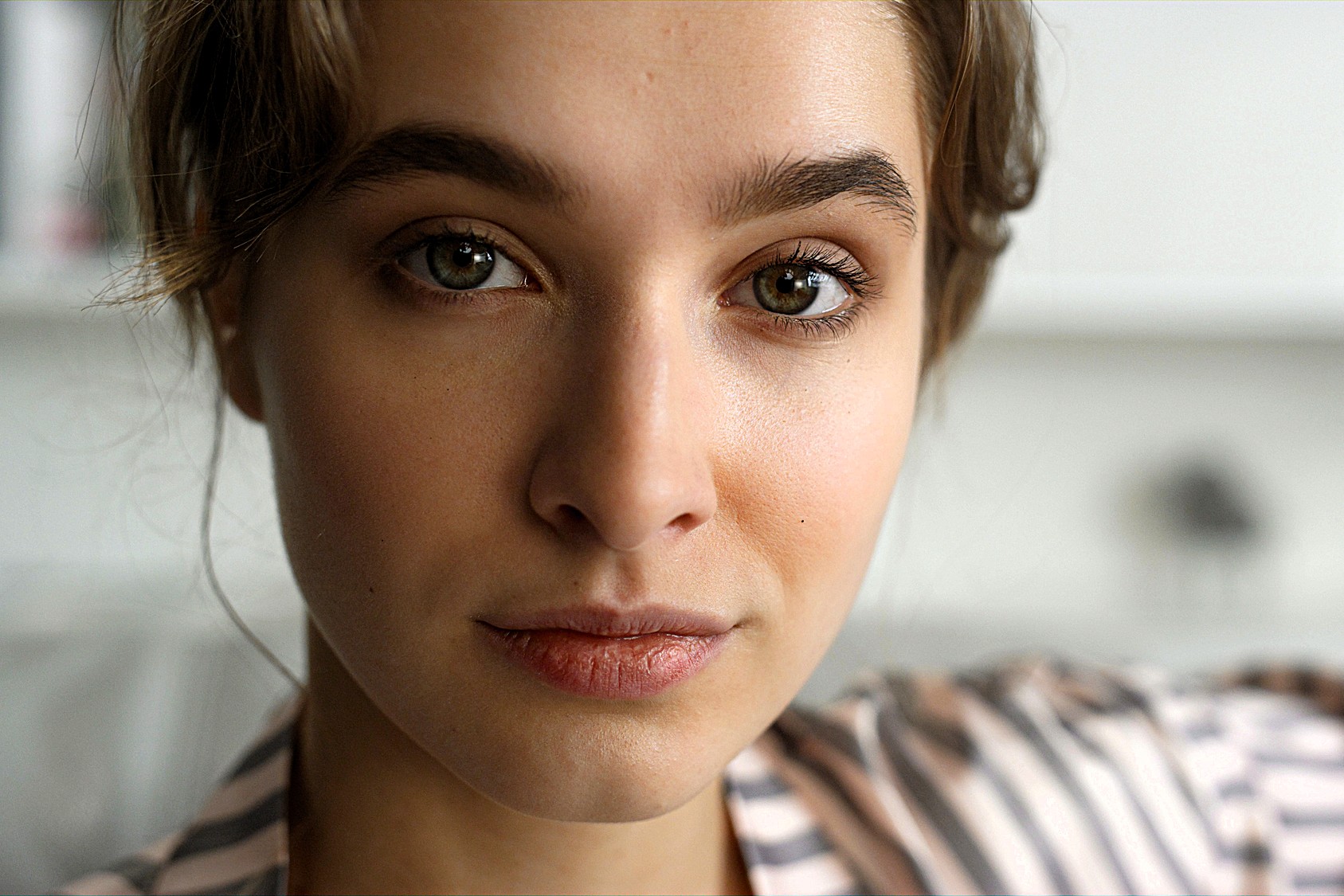 A young person with light brown hair and hazel eyes looking at the camera. They are wearing a striped shirt, and the background is blurred.