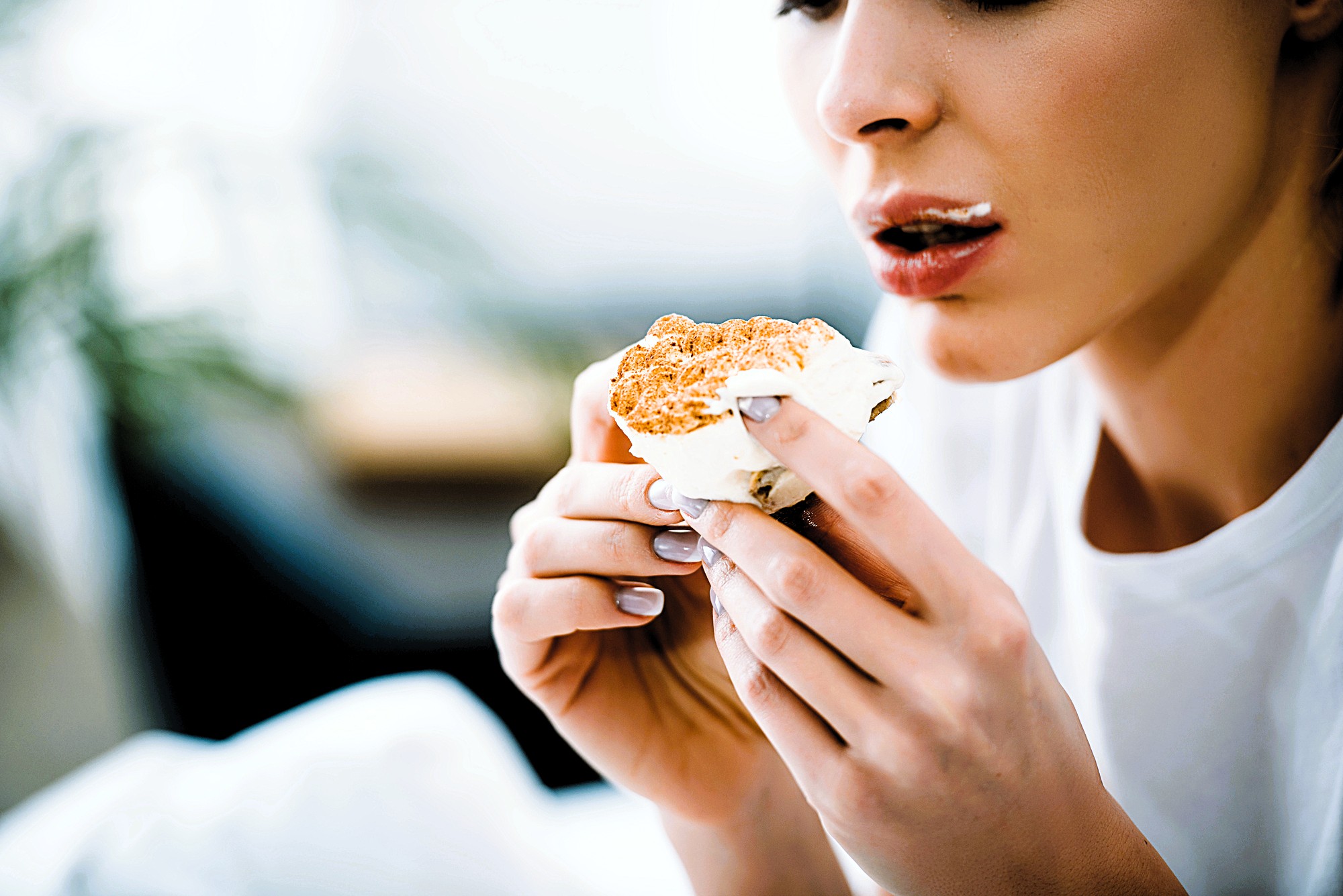 Person holding a cupcake with frosting, bringing it to their mouth. Some frosting is smudged on their lips. They are wearing a white shirt, and the background is softly blurred, indicating an indoor setting.