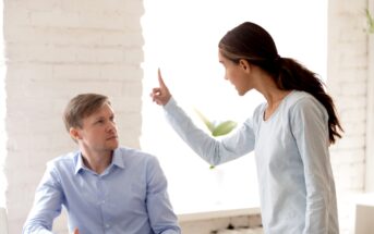 A woman with long hair wearing a light blue top is standing and pointing her finger while speaking to a seated man in a blue shirt. The scene appears to be indoors with white brick walls and bright lighting.
