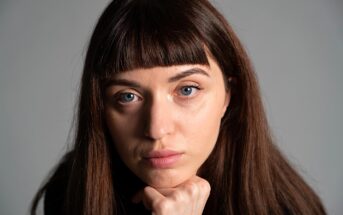 A person with long brown hair and bangs looks directly at the camera with a neutral expression, resting their chin on a fist. The background is a plain, light gray.