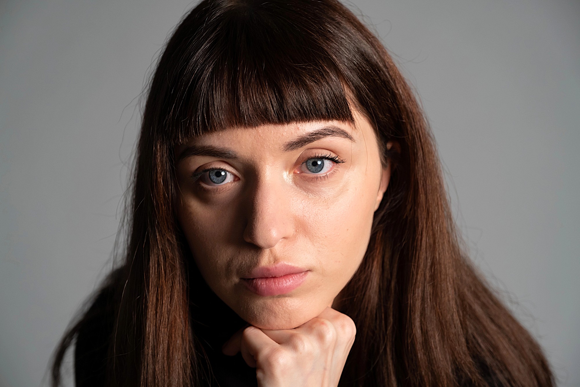 A person with long brown hair and bangs looks directly at the camera with a neutral expression, resting their chin on a fist. The background is a plain, light gray.