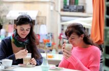 Two women are sitting at an outdoor café. One woman in a pink sweater sips from a tall glass, while the other in a scarf stirs her drink. They are surrounded by tables and chairs, with buildings and umbrellas in the background.