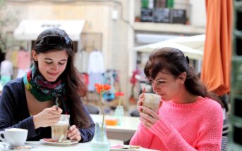 Two women are sitting at an outdoor café. One woman in a pink sweater sips from a tall glass, while the other in a scarf stirs her drink. They are surrounded by tables and chairs, with buildings and umbrellas in the background.