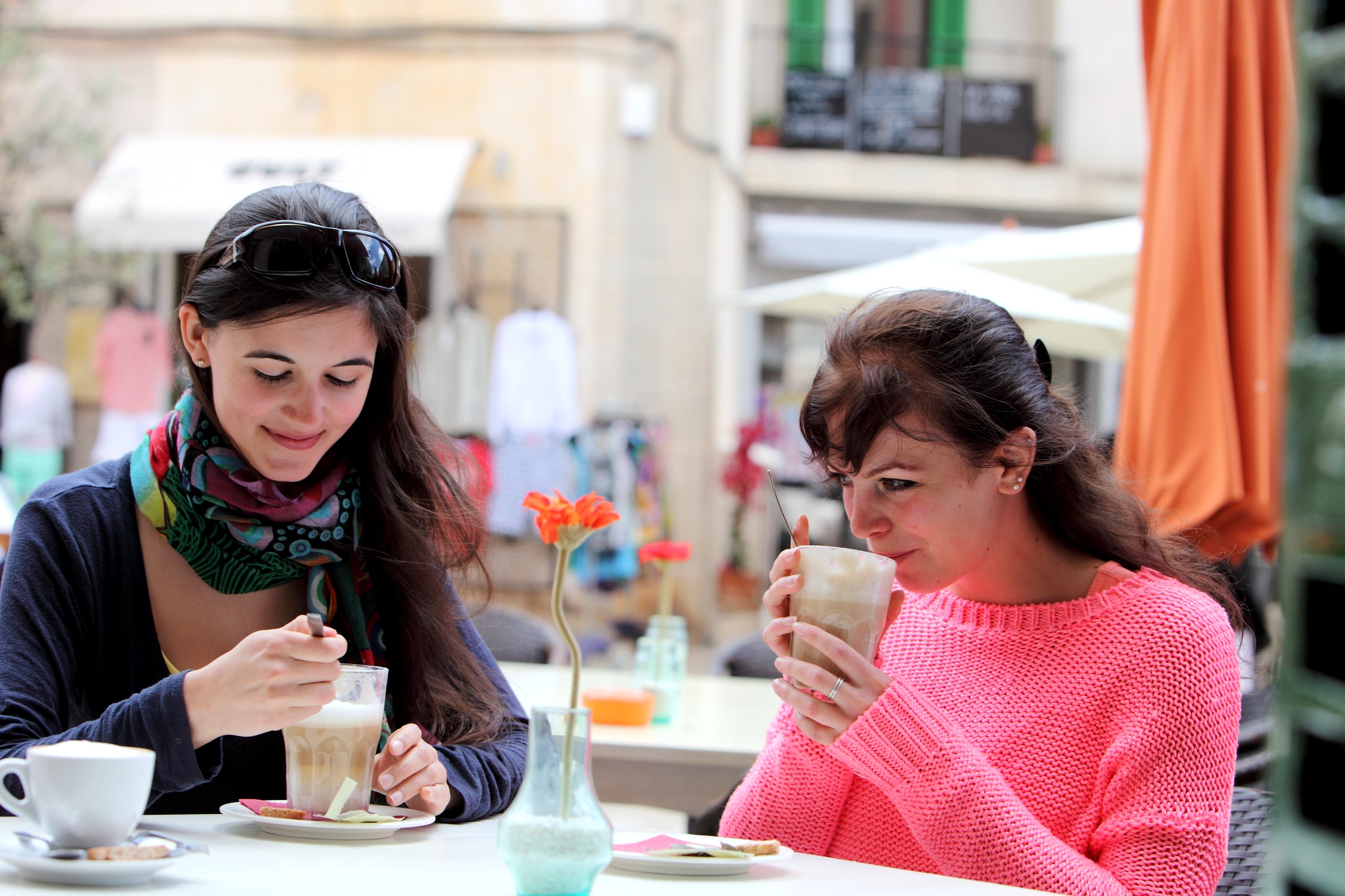 Two women are sitting at an outdoor café. One woman in a pink sweater sips from a tall glass, while the other in a scarf stirs her drink. They are surrounded by tables and chairs, with buildings and umbrellas in the background.