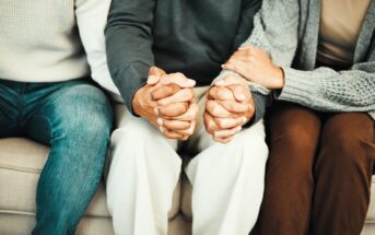 Three people sit closely on a couch, holding hands. They wear casual, cozy sweaters and jeans. Their intertwined hands suggest support and unity, while their bodies are partially visible, conveying a sense of togetherness.
