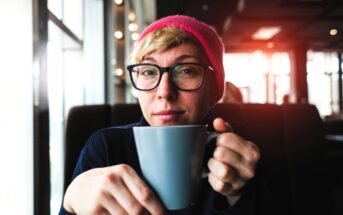 Person wearing glasses and a pink beanie, holding a gray mug, sits in a cozy cafe with sunlight streaming in through the windows.