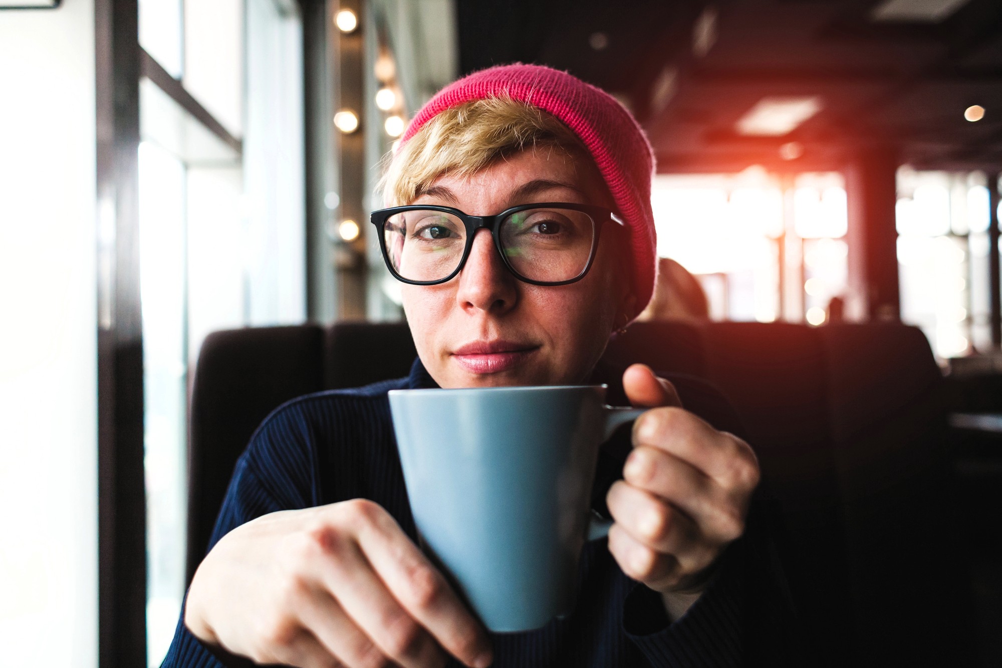 Person wearing glasses and a pink beanie, holding a gray mug, sits in a cozy cafe with sunlight streaming in through the windows.