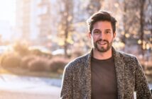 A man with short hair and a beard smiles while standing outdoors in a sunlit, blurry urban setting. He is wearing a gray sweater over a dark shirt.