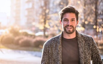 A man with short hair and a beard smiles while standing outdoors in a sunlit, blurry urban setting. He is wearing a gray sweater over a dark shirt.