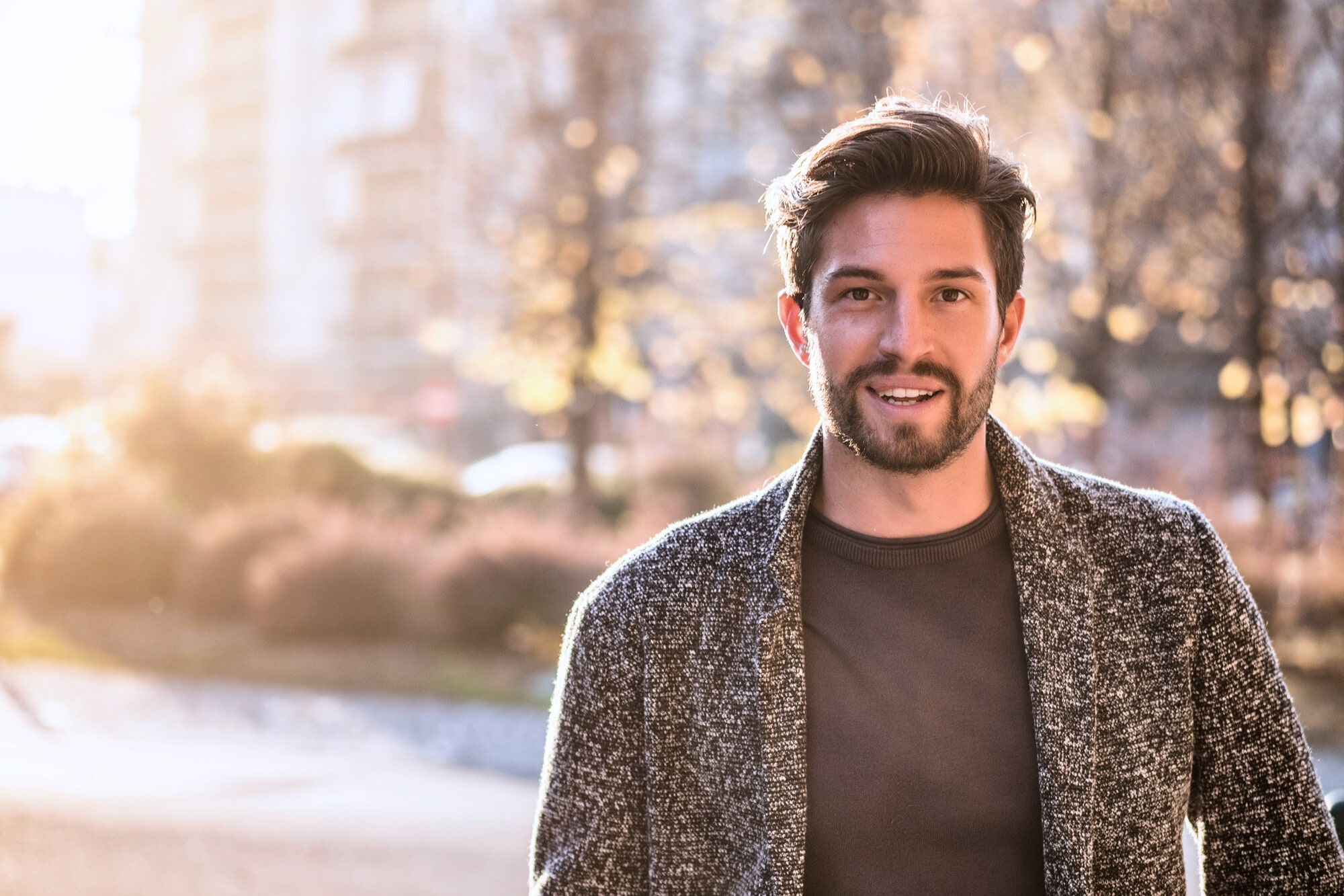 A man with short hair and a beard smiles while standing outdoors in a sunlit, blurry urban setting. He is wearing a gray sweater over a dark shirt.