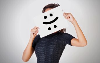 A person in a dark shirt holds a piece of paper in front of their face. The paper has a smiley face drawn with eyes and a crooked line for a mouth, resembling a puzzled or mixed expression. The background is plain and light gray.