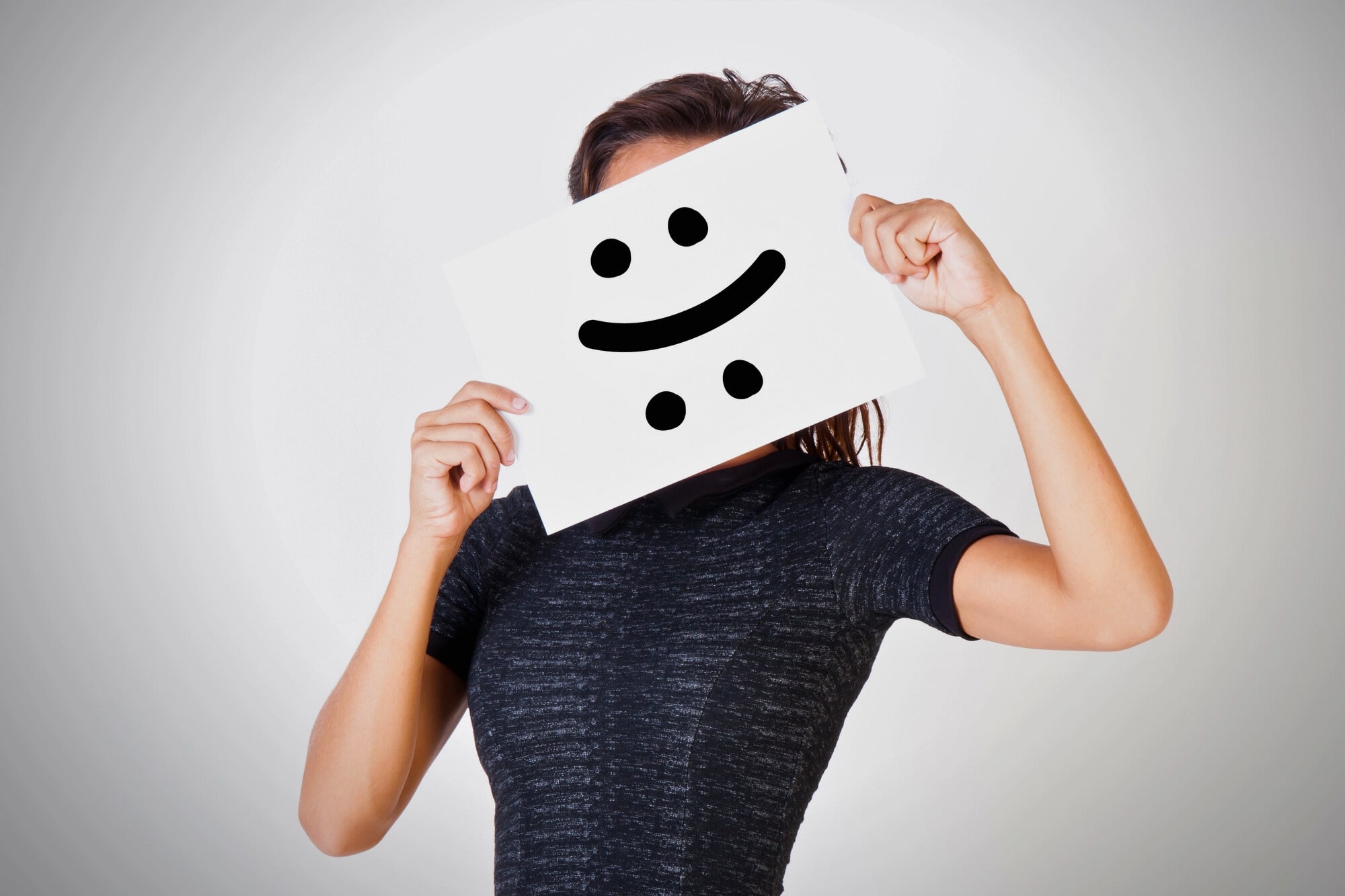 A person in a dark shirt holds a piece of paper in front of their face. The paper has a smiley face drawn with eyes and a crooked line for a mouth, resembling a puzzled or mixed expression. The background is plain and light gray.