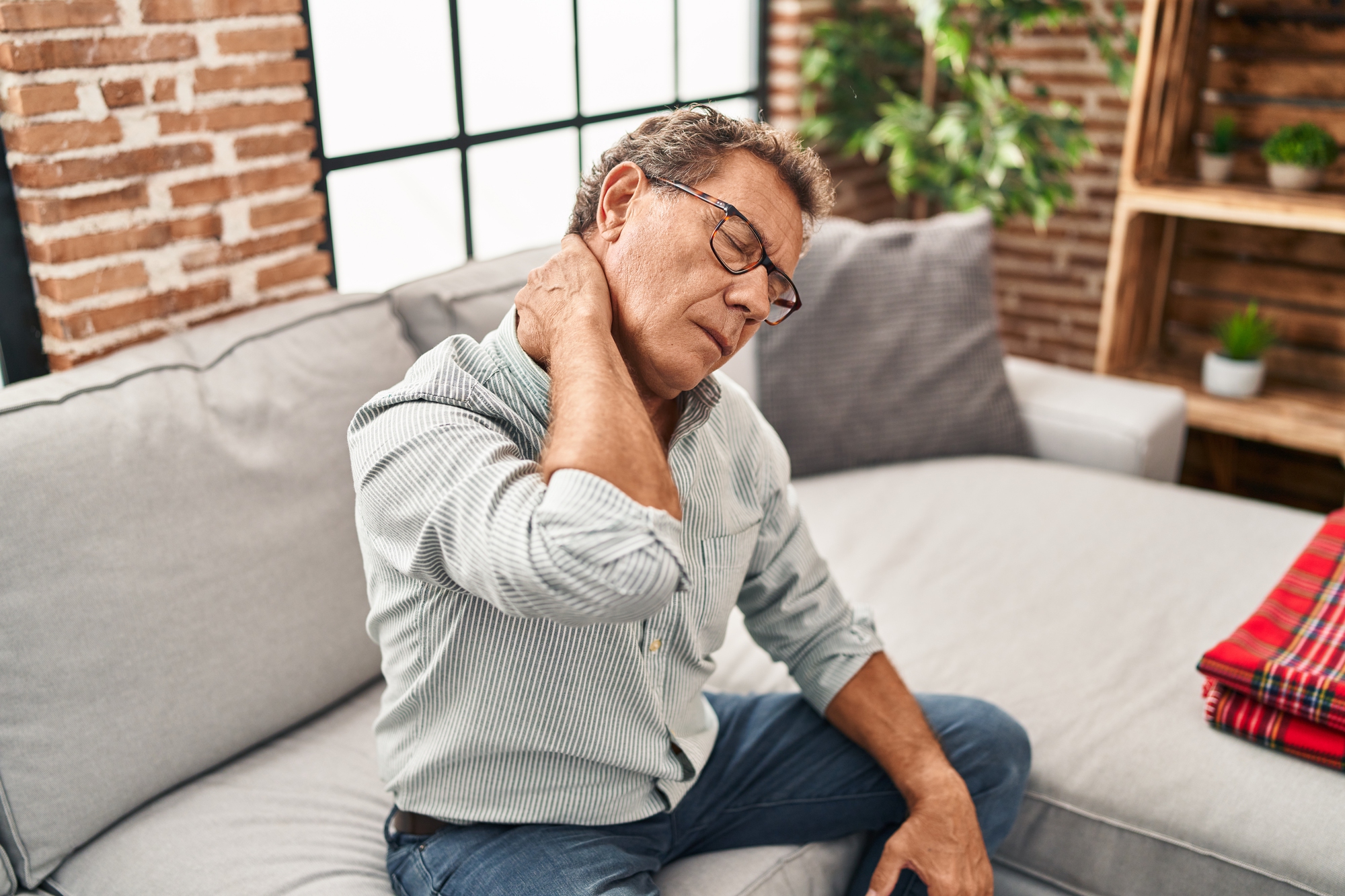 Man sitting on a gray couch with one hand on his neck, appearing to have neck pain. He is wearing glasses and a striped shirt. The room has brick walls and plants, with a shelf in the background.