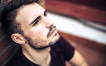 A man with short hair and a beard is leaning against a brick wall. He is gazing upwards with a focused and contemplative expression. The background is blurred, emphasizing the subject's face.
