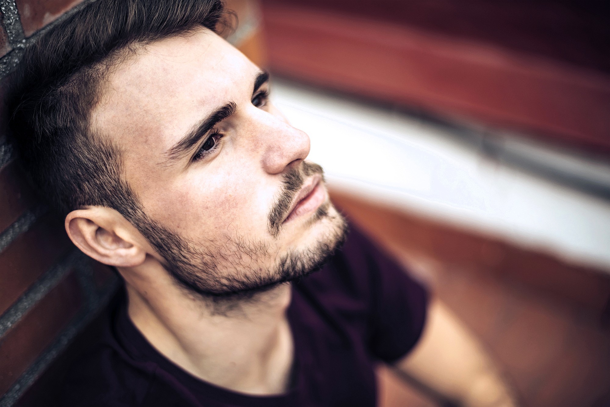 A man with short hair and a beard is leaning against a brick wall. He is gazing upwards with a focused and contemplative expression. The background is blurred, emphasizing the subject's face.