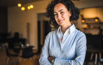 Woman with curly hair and a white blouse smiles confidently while standing with arms crossed in a warmly lit room. The background includes blurred furnishings and soft lighting.