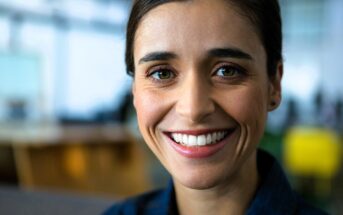 A person with dark hair smiles warmly, wearing a denim shirt. The background is softly blurred, suggesting an indoor setting. The lighting highlights their face, giving the image a cheerful and welcoming vibe.